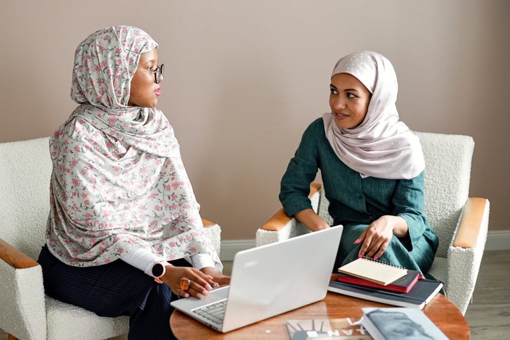 Two Muslim businesswomen engaging in a meeting at an office space with laptops and notes.